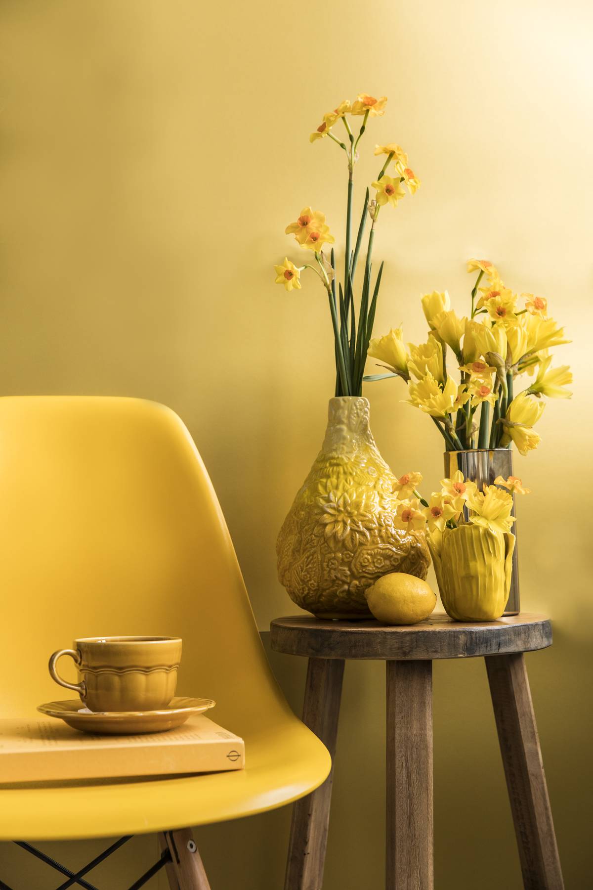 Gele stoel, kopje op boek, vaas met narcissen en citroen op houten krukje tegen gele achtergrond.