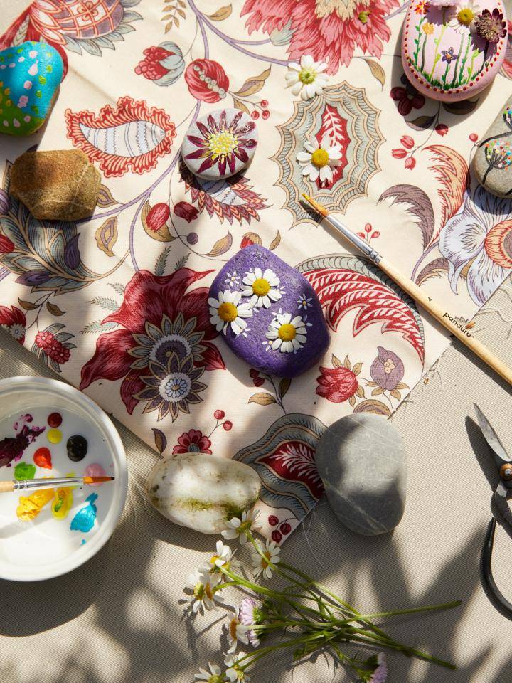 Twee mensen schilderen stenen met bloemenpatronen op een tafel bedekt met een bloemendoek.