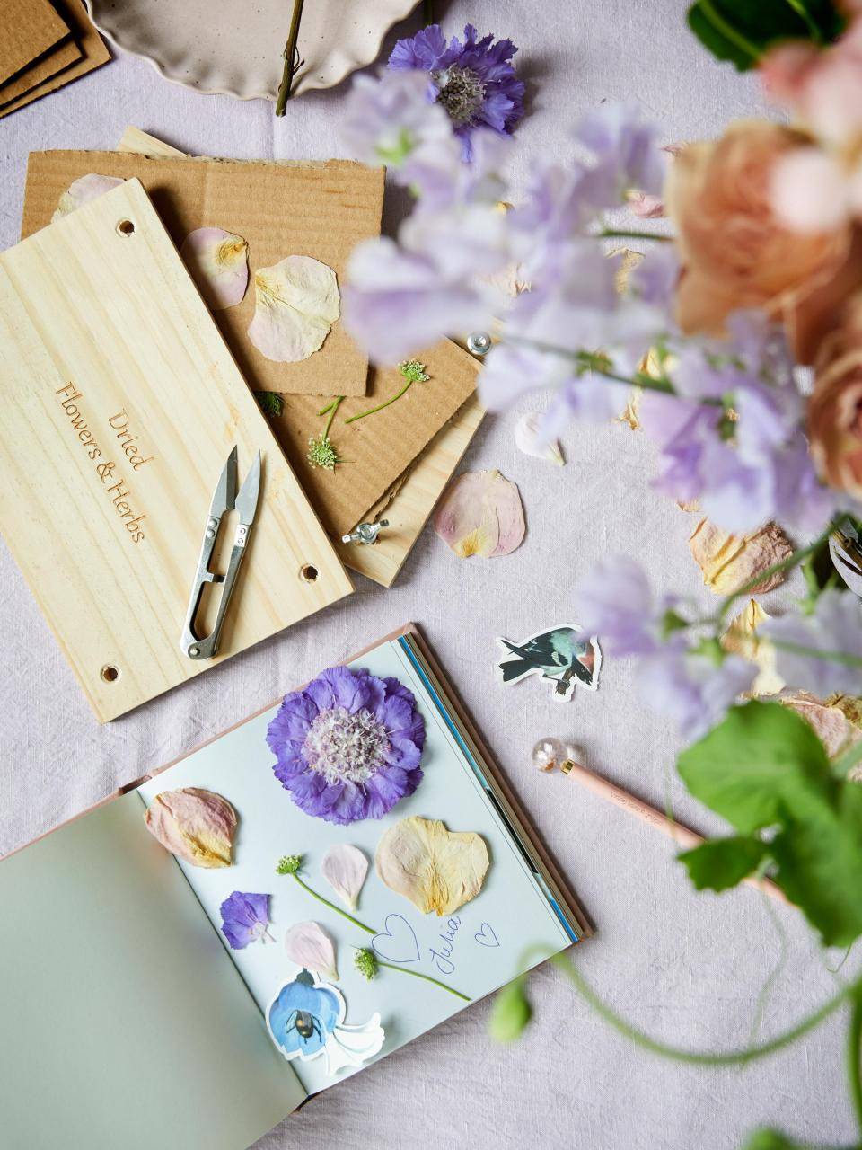 Open boek met gedroogde bloemen en handgeschreven notitie, omringd door bloemendecoraties en gereedschap.