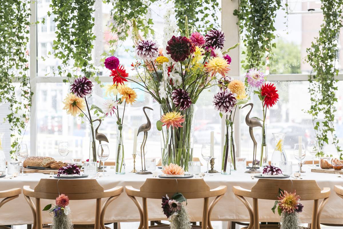 Sierlijk gedekte tafel met kleurrijke bloemen in hoge vazen, kraanvogelbeelden en groene klimop op de achtergrond.