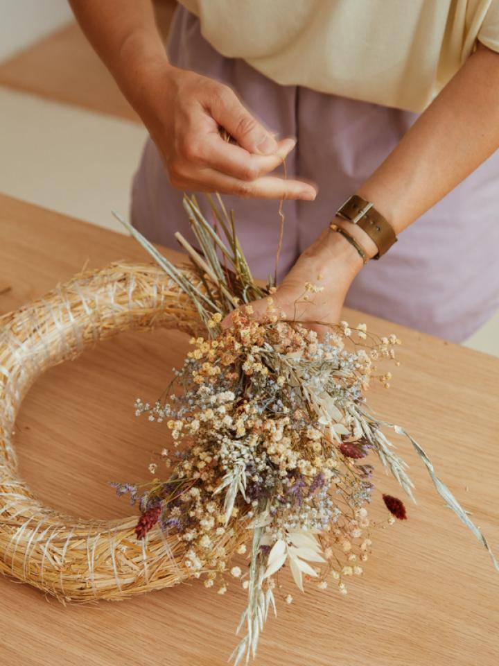 Person arranging dried flowers into a wreath on a wooden surface.