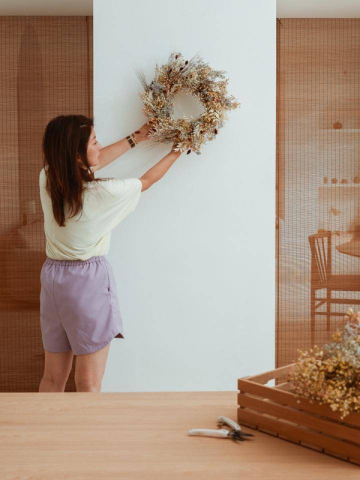 Person hanging a decorative wreath on a white wall, with dried flowers and tools on a wooden table.