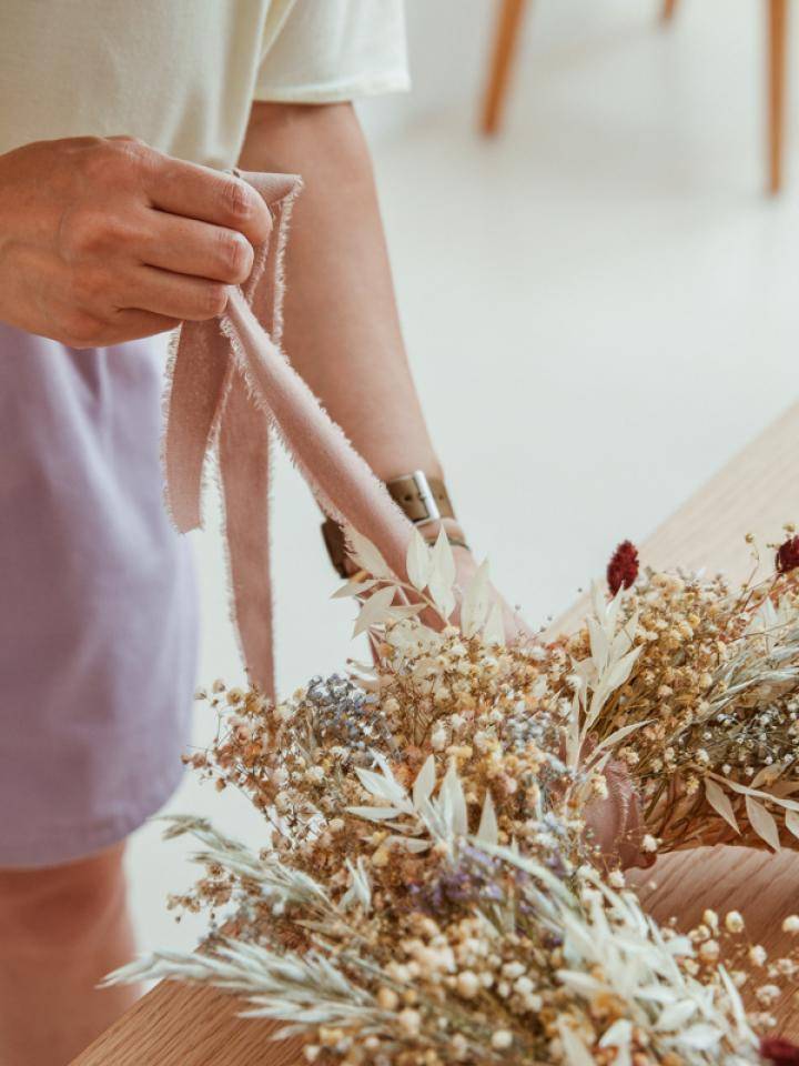 Person arranging dried flowers on a wooden surface, holding pink fabric to tie them together.