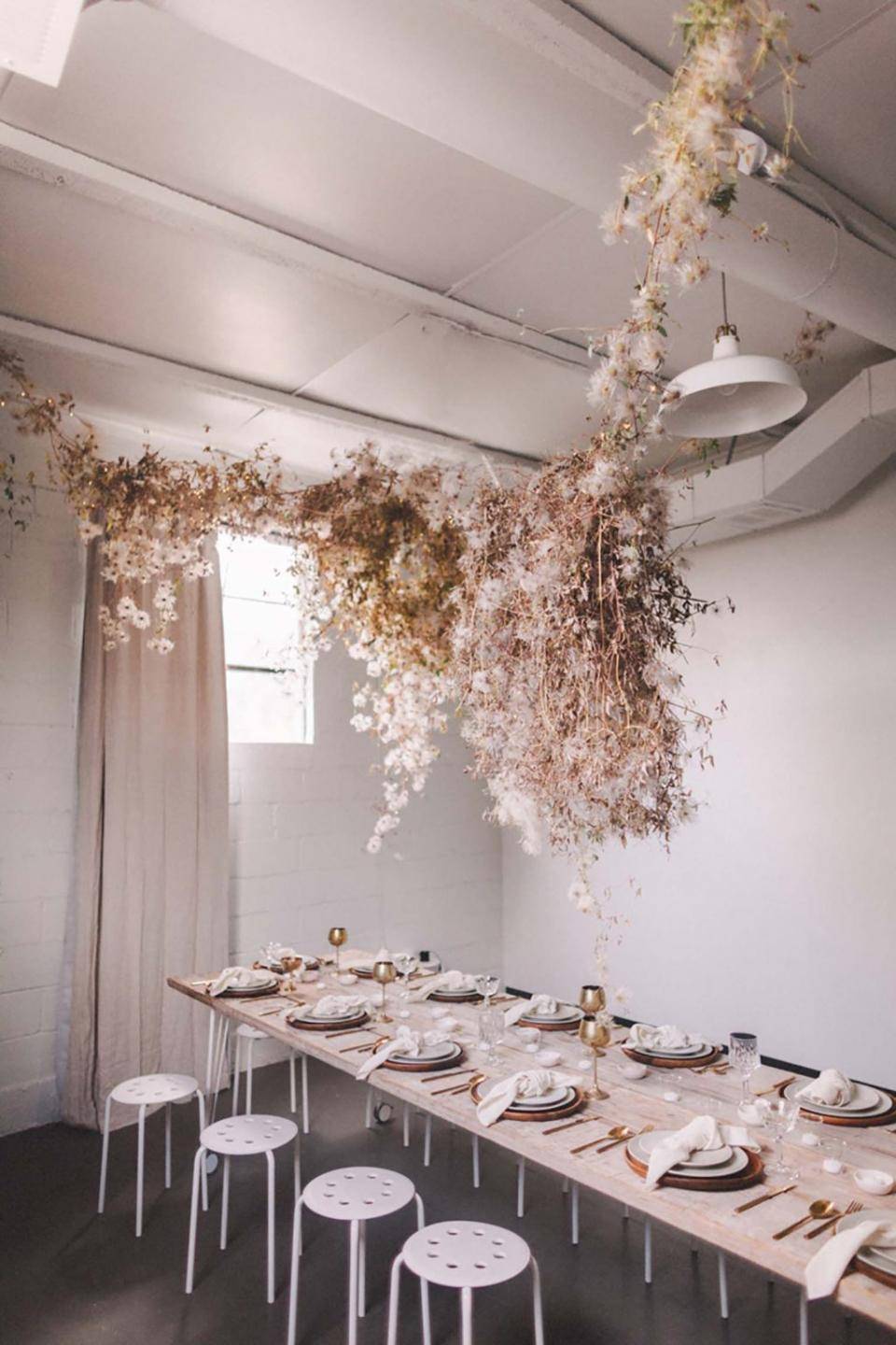 Eettafel gedekt voor een maaltijd, met borden, bestek en glazen, en een hangend bloemstuk van gedroogde bloemen en groen.