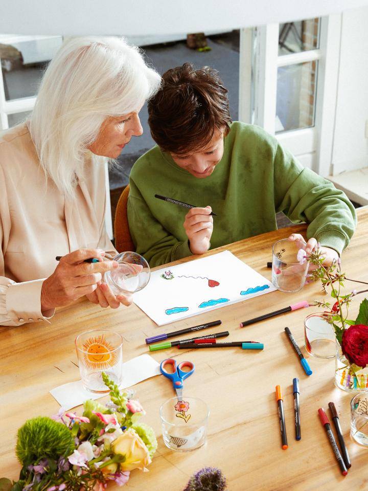 : Twee mensen aan een tafel bezig met een kunstactiviteit, met bloemen, stiften en glazen op de tafel.