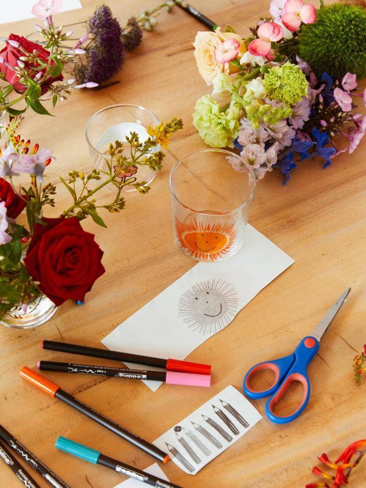 Bloemen, stiften, schaar en een glas op een houten tafel met een tekening van een lachend gezicht.