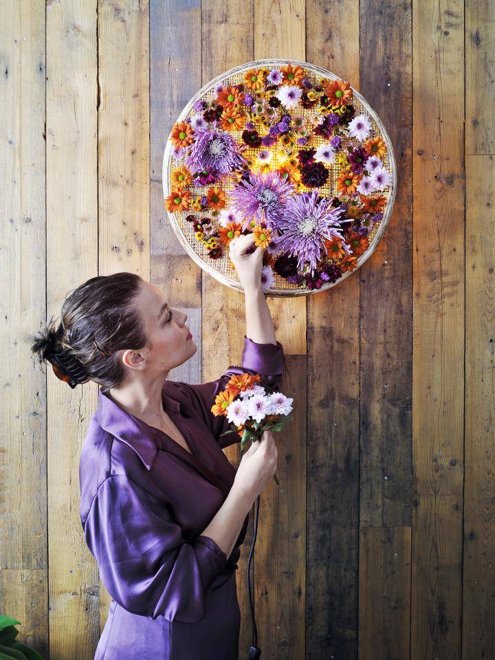 Vrouw in paarse jurk hangt bloemenkunstwerk aan houten muur.