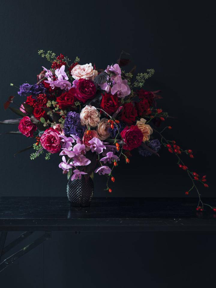Kerstboeket met rode, paarse en roze bloemen in een zwarte vaas tegen een donkere achtergrond.