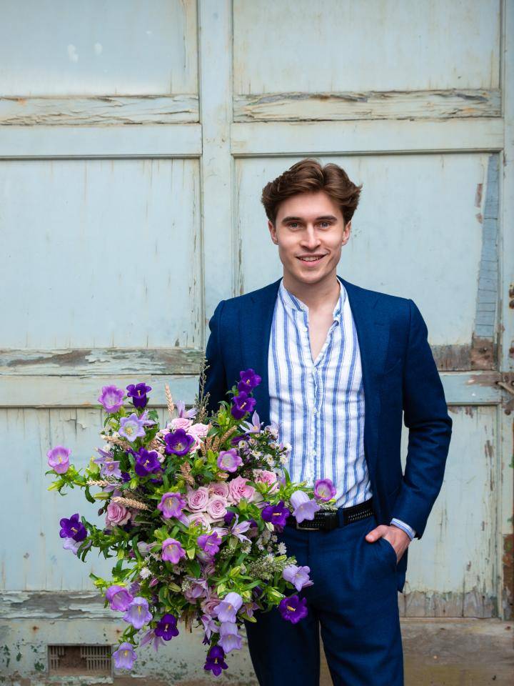 Persoon in blauw pak met gestreept overhemd, houdt een groot boeket van paarse en roze bloemen vast, voor een verweerde houten deur.
