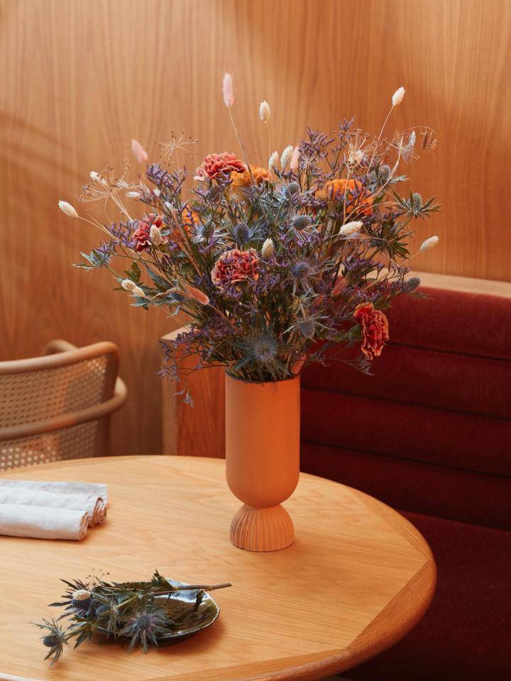 Boeket van distels en droogbloemen in een vaas op een houten tafel met stoel en bank op de achtergrond.